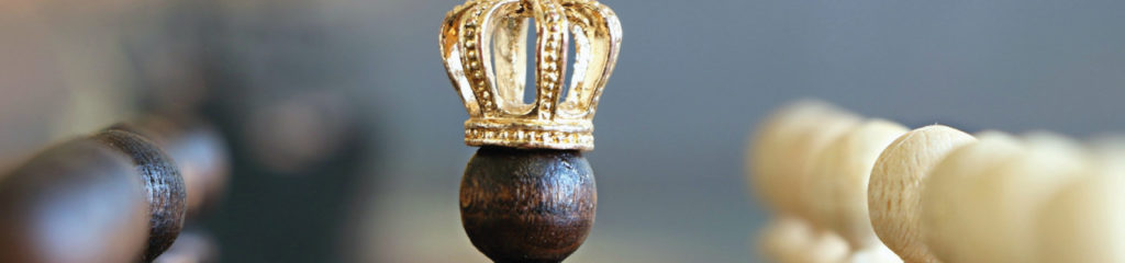 imagem de um peão de xadrez com uma coroa. Peças brancas e pretas em seu redor como se esse peão estivesse passando em meio aos seus suditos.