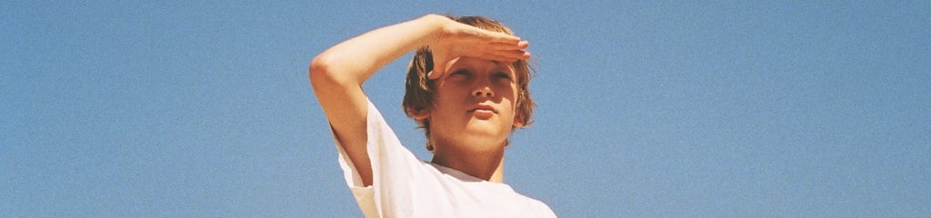 Imagem de um menino, de cabelos loiros, com a mão no rosto, tapando o sol. Ele olha para frente.