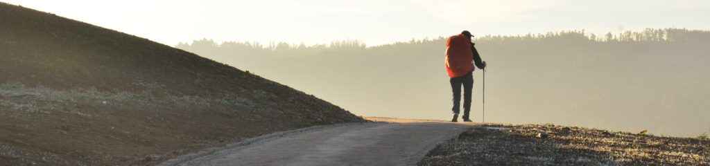 Imagem de uma pessoa caminhando por um campo vazio. Ela carrega uma mochila vermelha nas costas.